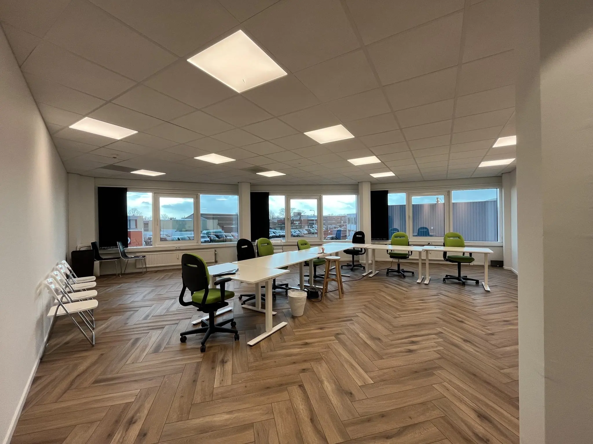 Vergaderruimte aan de Lange Dreef met moderne bureaustoelen, houten visgraatvloer en grote ramen die uitzicht bieden op parkeerplaats en gebouwen.